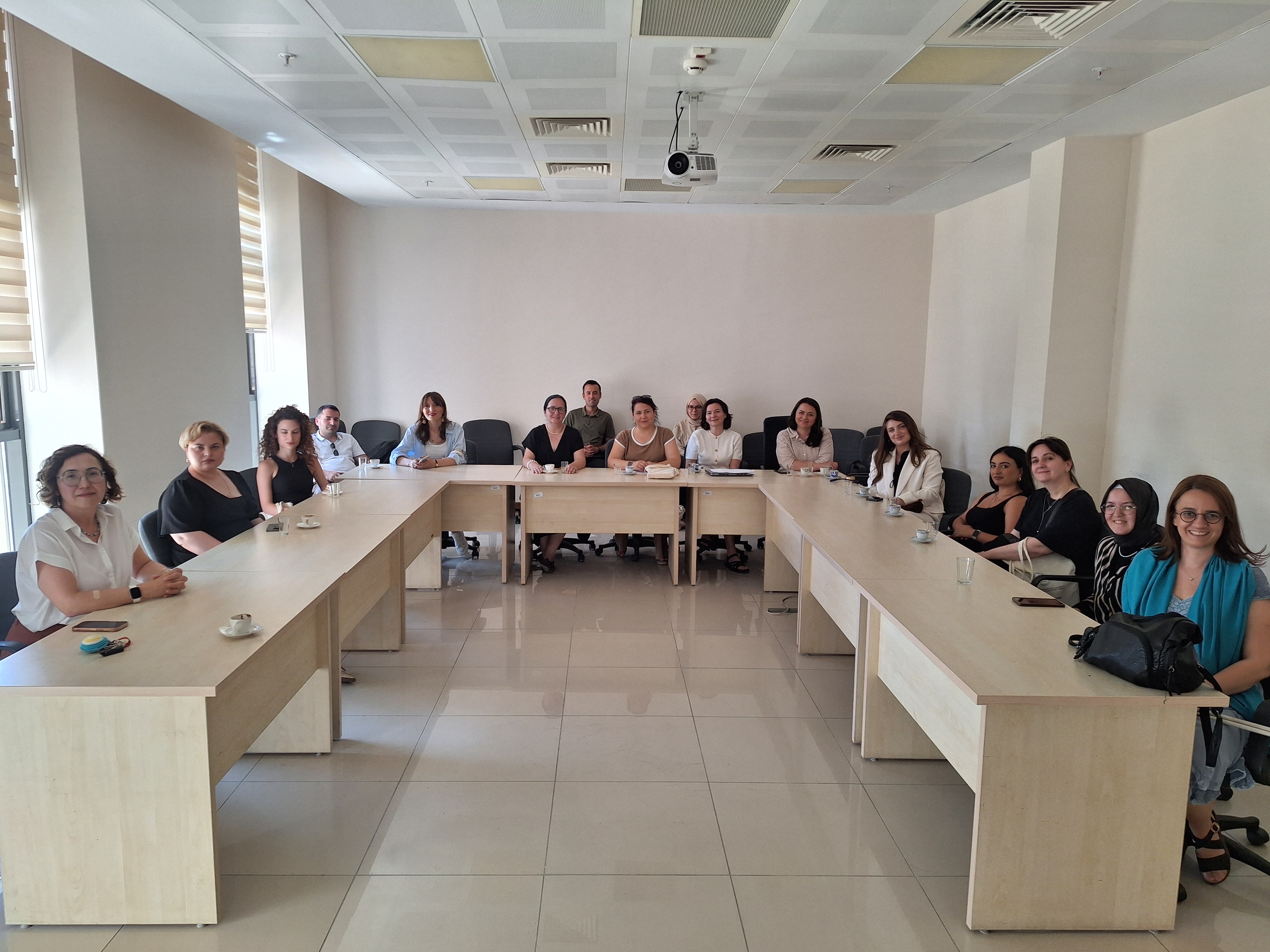 Aydınlık bir toplantı salonunda, U düzenindeki masanın etrafında oturan çoğunluğu kadın bir grup kameraya gülümsüyor. Modern ofis ortamındaki ekip, profesyonel bir atmosferde, masadaki kahve fincanları eşliğinde bir arada poz veriyor.