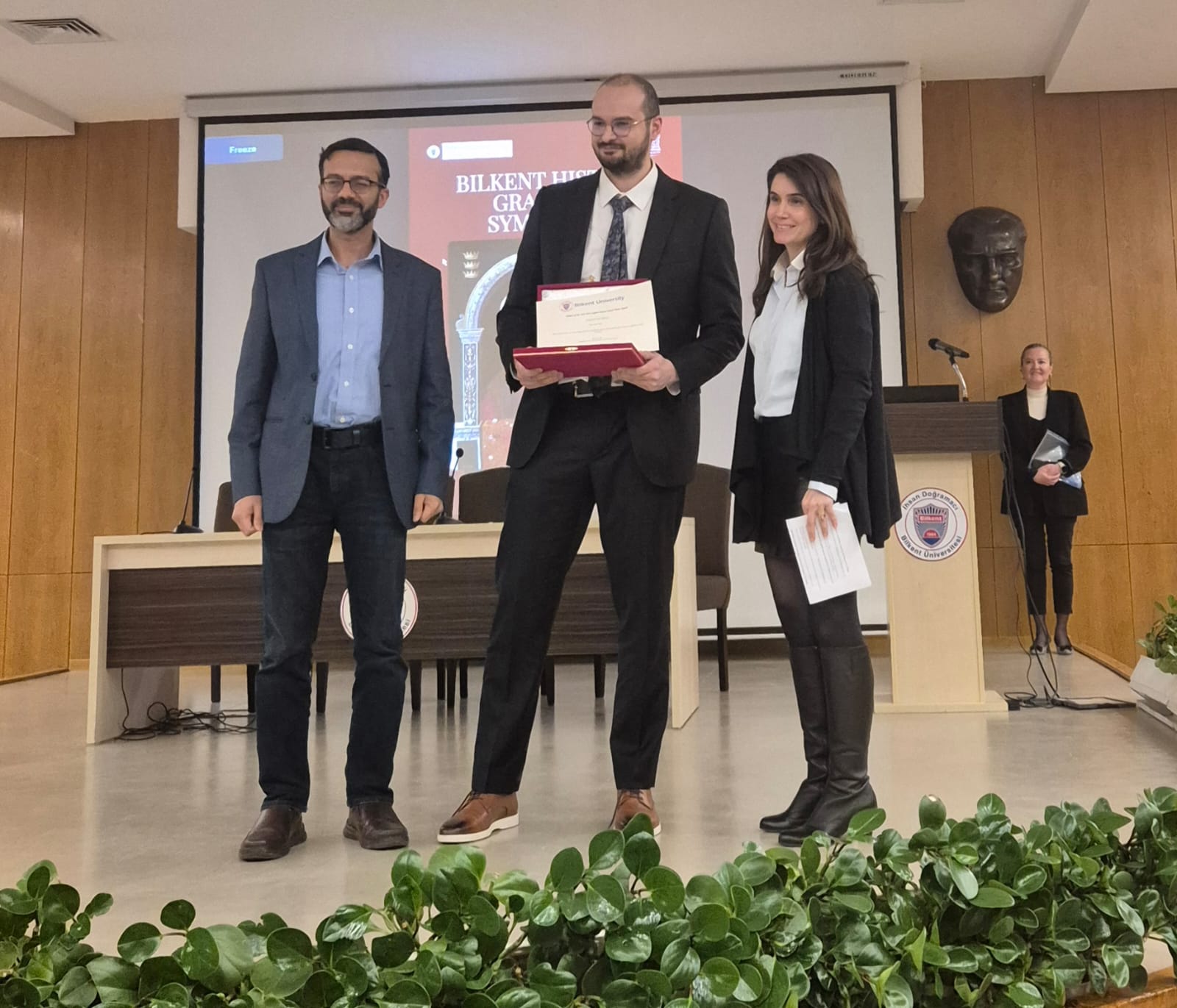 TR: Hasan Duymuş (ortada), Bilkent Üniversitesi'nde Çiğdem Barçan Tansel En İyi Yüksek Lisans Tezi Ödülü'nü alırken iki öğretim üyesi ile sahnede, elinde ödül beratıyla gülümsüyor. EN: Hasan Duymuş (center), holding his award certificate, smiles with two faculty members on stage at Bilkent University while receiving the Çiğdem Barçan Tansel Best Master's Thesis Award.